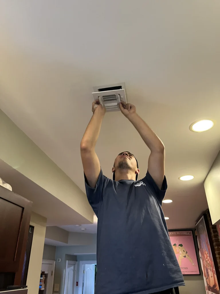 A person in a blue shirt adjusts a ceiling vent in a well-lit room with recessed lighting and modern decor.